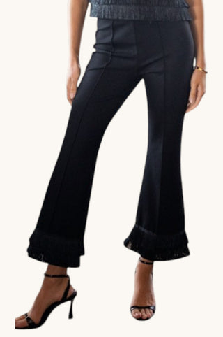 Black flared pants with fringe detailing at the hem, shown on a model wearing black strappy high-heeled sandals against a plain off-white background.
