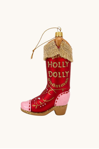 A Christmas ornament shaped like a cowboy boot, featuring a red shaft with gold glittery lettering that reads 'HOLLY DOLLY Christmas', a light pink foot, a beige fringed collar, and a gold hanging loop, shown on a plain off-white background.
