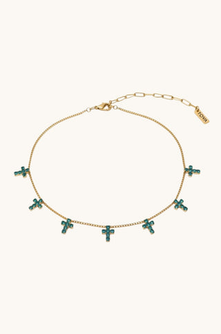 A gold chain necklace featuring multiple small cross charms embellished with green crystals, displayed on a light cream background. The brand name 'MIMI' is visible on a small gold tag near the clasp.