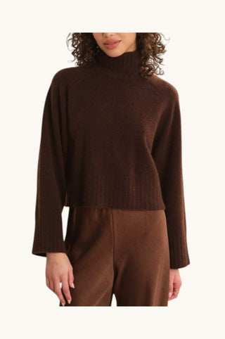 A woman wearing a dark brown ribbed knit mock-neck sweater and matching brown trousers, shown from the mouth down against a plain off-white background.