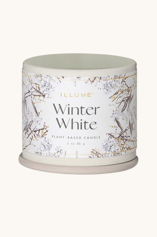 A cylindrical plant-based candle tin by ILLUME, labeled 'Winter White,' with a white label decorated with delicate gray and gold floral/berry branch illustrations, shown on a light cream background.