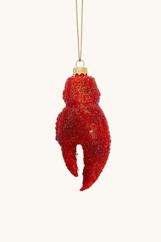 A red lobster claw Christmas ornament covered in textured glitter, hanging from a gold string against a solid off-white background.
