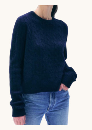 A person wearing a dark navy blue cable-knit crewneck sweater and blue jeans, shown from the neck down against a plain off-white background.