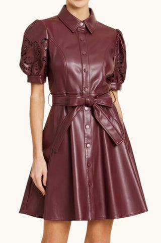 A woman wearing a short-sleeve faux-leather dress in deep burgundy with floral lace applique on the sleeves, a pointed collar, front buttons, and a self-tie belt at the waist, shown against a plain light background.