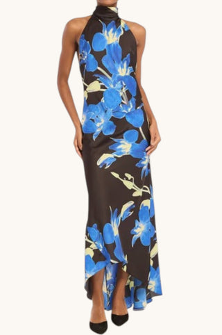 A woman modeling a sleeveless halter-neck maxi dress with a high-low hem. The dress features a dark brown base with a vibrant blue and light yellow floral print, set against a plain off-white background.