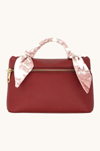 A burgundy pebbled leather handbag with a top zipper, gold-tone hardware, and a pink and white patterned silk scarf tied to the handle, shown against a plain off-white background.