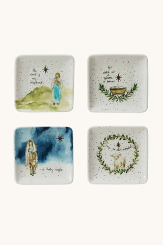 Four small square white ceramic plates with watercolor Christmas illustrations, arranged in a 2x2 grid on a light cream background. The designs include a shepherd by a green hill, a manger scene with a gold bowl, a shepherd under a blue night sky, and a lamb framed by a green wreath.