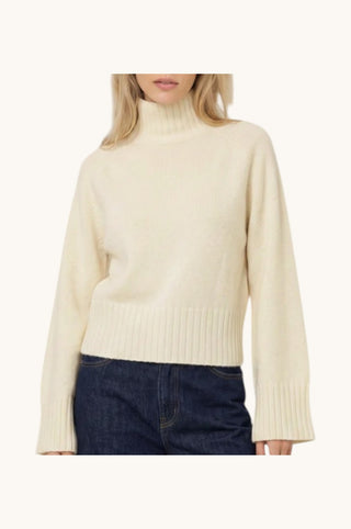 A woman wearing a cream-colored ribbed turtleneck sweater and dark blue jeans, shown from the neck down against a solid off-white background.