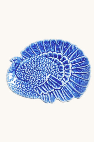 A blue and white glazed ceramic plate shaped like a turkey, featuring a textured, raised design on the body and tail feathers, set against a plain off-white background.