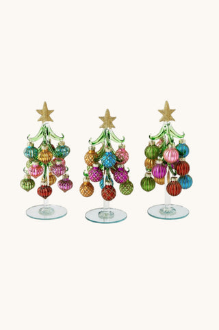 Three small glass Christmas tree ornaments with gold star toppers and colorful hanging baubles on clear stands, shown against a plain light beige background.