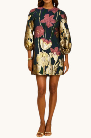 A woman modeling a short puff-sleeve dress with a dark navy background and a large metallic gold and dusty pink floral print, set against a plain off-white background.