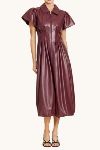 A model wearing a short-sleeved, collared faux-leather midi dress in a deep burgundy color, paired with black strappy heels, against a plain off-white background.